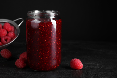 Photo of Delicious jam and fresh raspberries on black slate table, closeup. Space for text