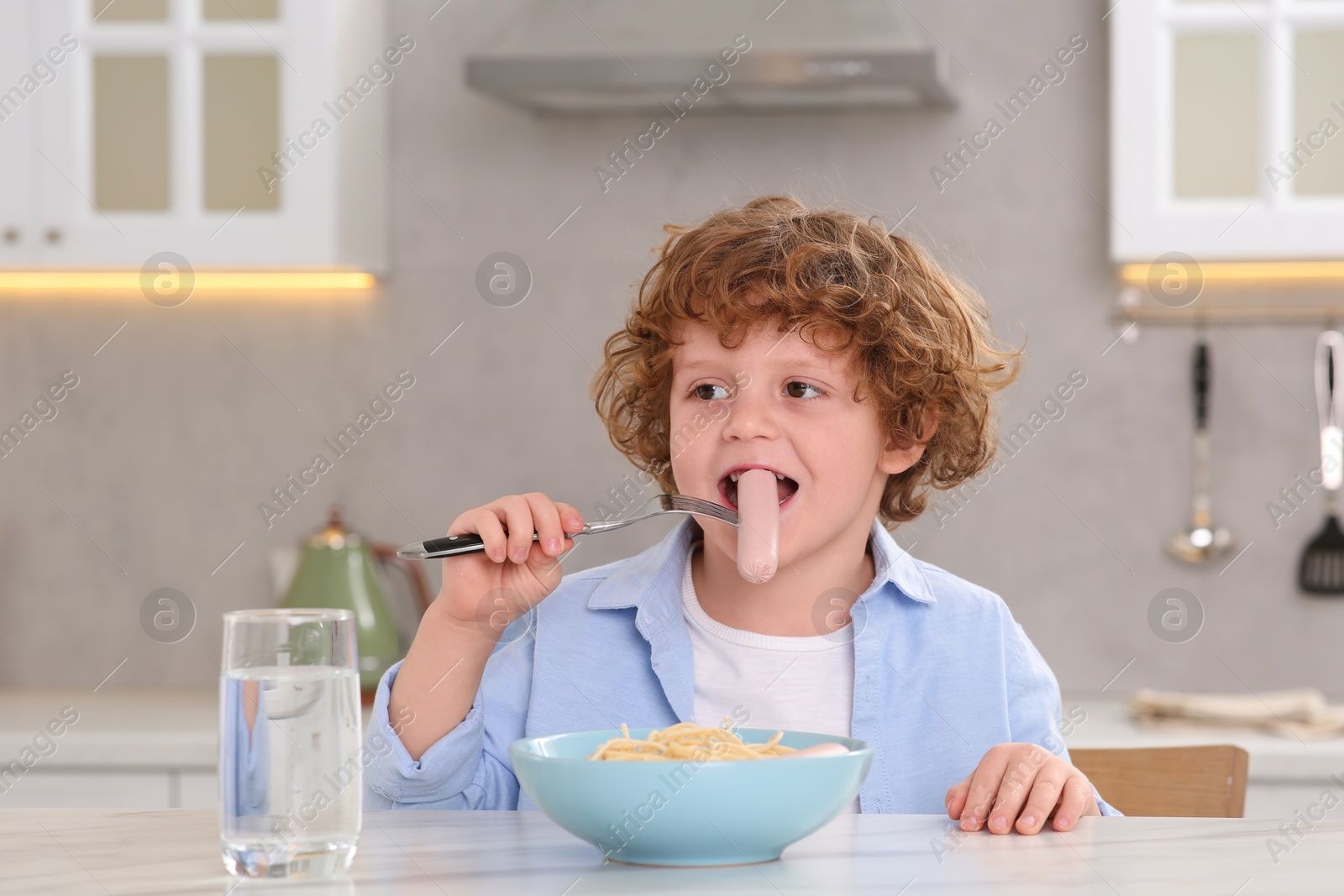 Photo of Cute little boy eating sausage and pasta at table in kitchen