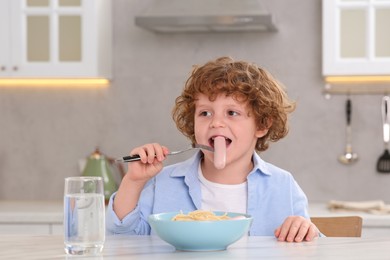 Cute little boy eating sausage and pasta at table in kitchen