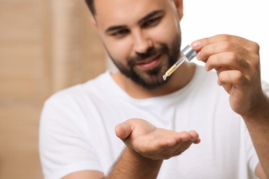 Young man applying cosmetic serum onto his hand indoors, selective focus