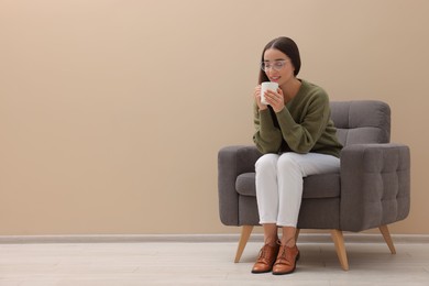 Photo of Beautiful woman with cup of drink sitting in armchair near beige wall indoors, space for text