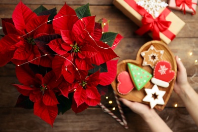 Woman with cookies near poinsettia (traditional Christmas flower) at wooden table, top view