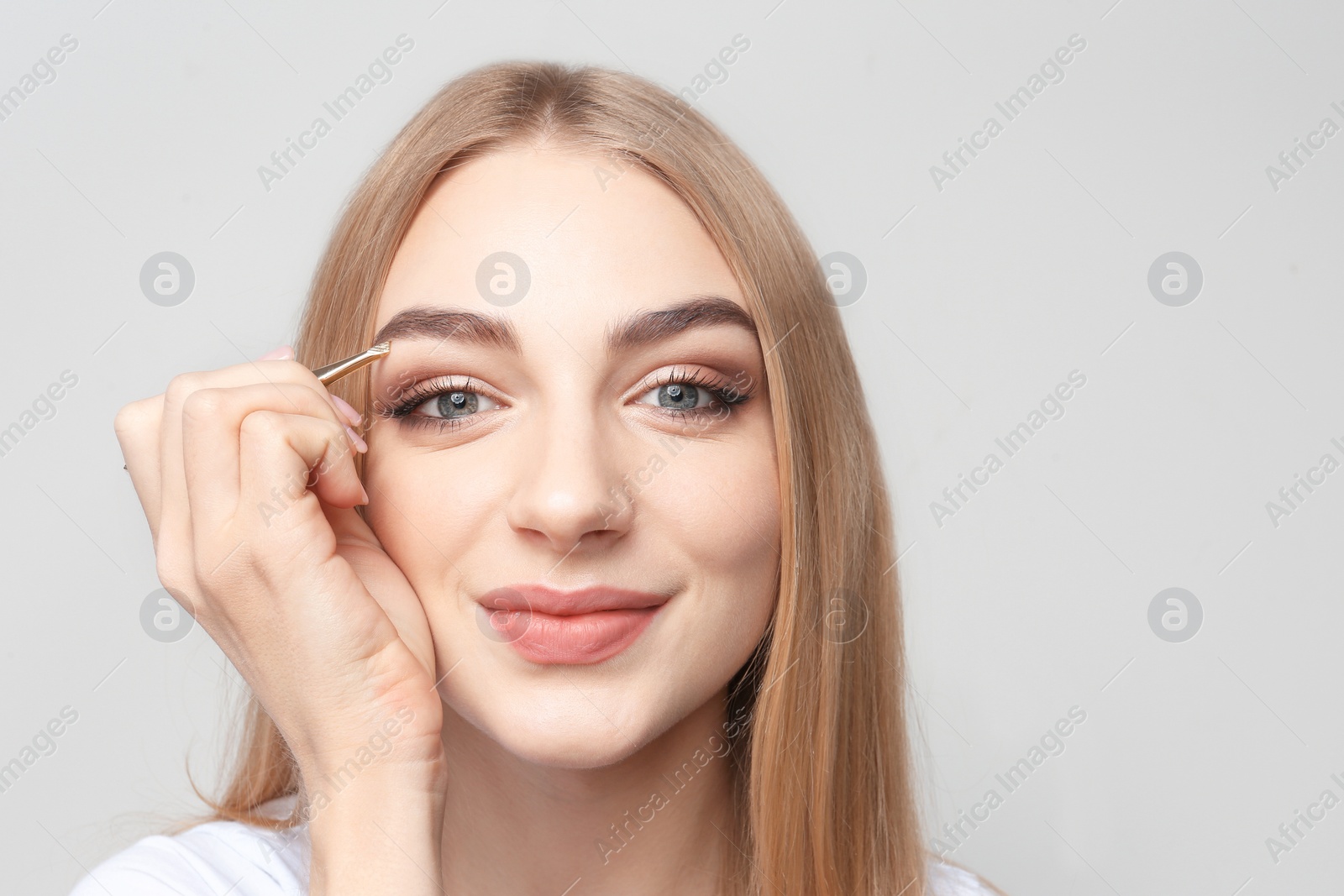 Photo of Young woman plucking eyebrow with tweezers on light background