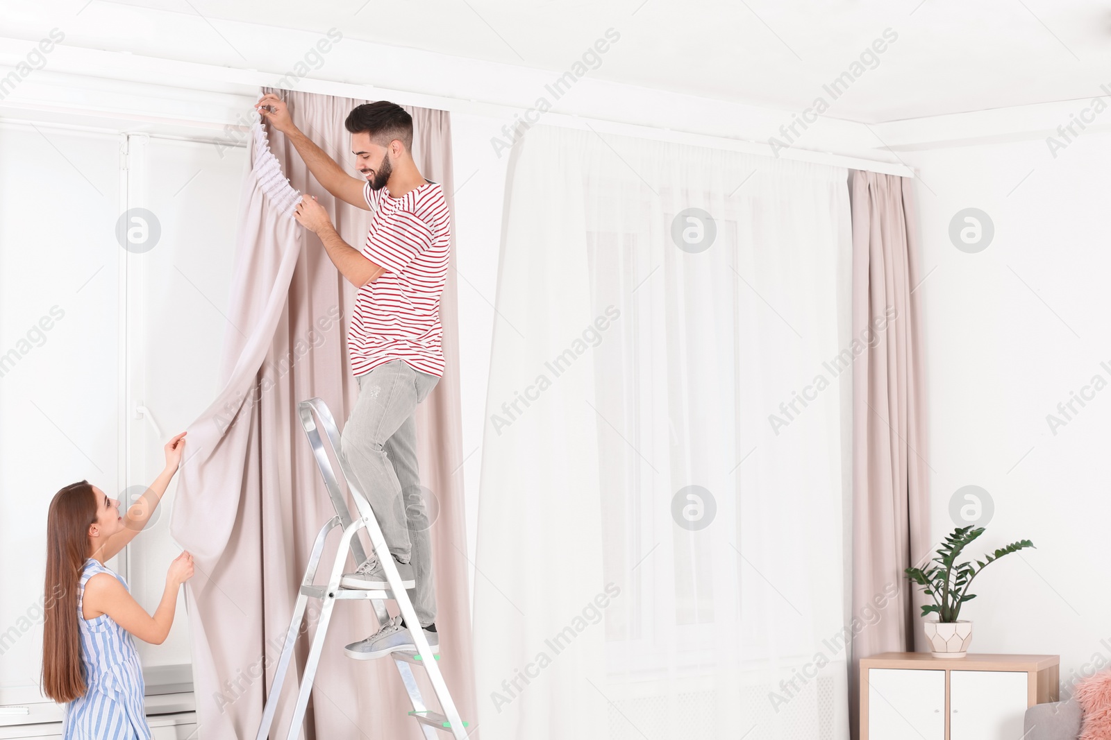 Photo of Young couple hanging window curtain in room. Space for text