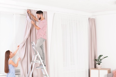 Young couple hanging window curtain in room. Space for text