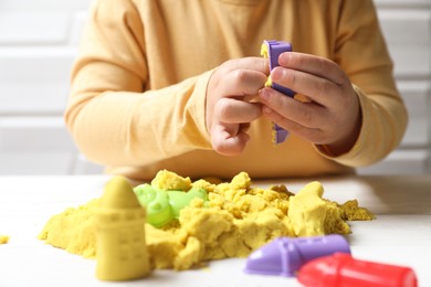 Photo of Little child playing with yellow kinetic sand at white table, closeup
