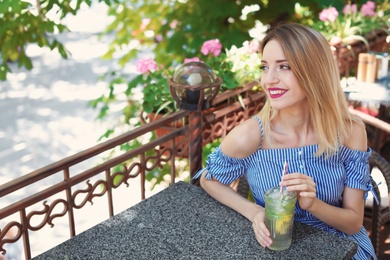 Photo of Young woman with glass of tasty lemonade at table in cafe, outdoors. Natural detox drink