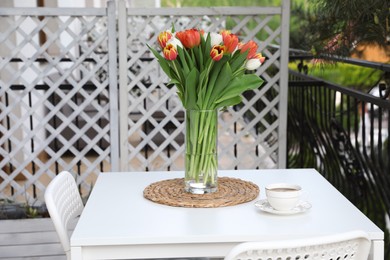 Beautiful colorful tulips in glass vase and cup of drink on white table at balcony