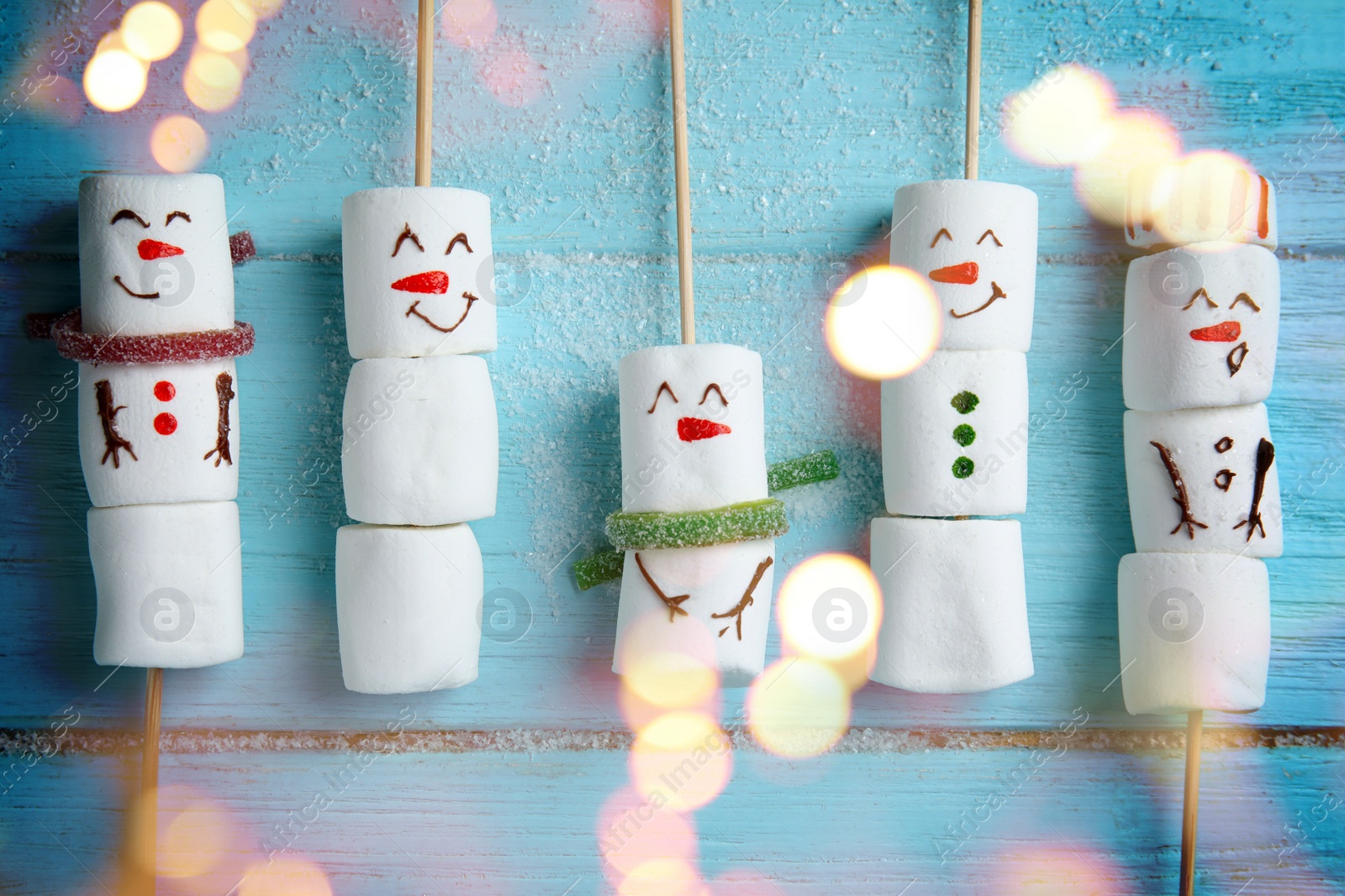 Image of Cute snowmen made of marshmallows on light blue wooden background, flat lay. Bokeh effect