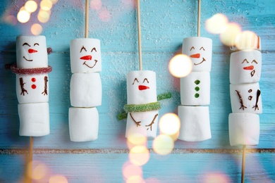 Image of Cute snowmen made of marshmallows on light blue wooden background, flat lay. Bokeh effect