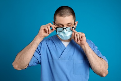 Photo of Doctor wiping foggy glasses caused by wearing disposable mask on blue background. Protective measure during coronavirus pandemic