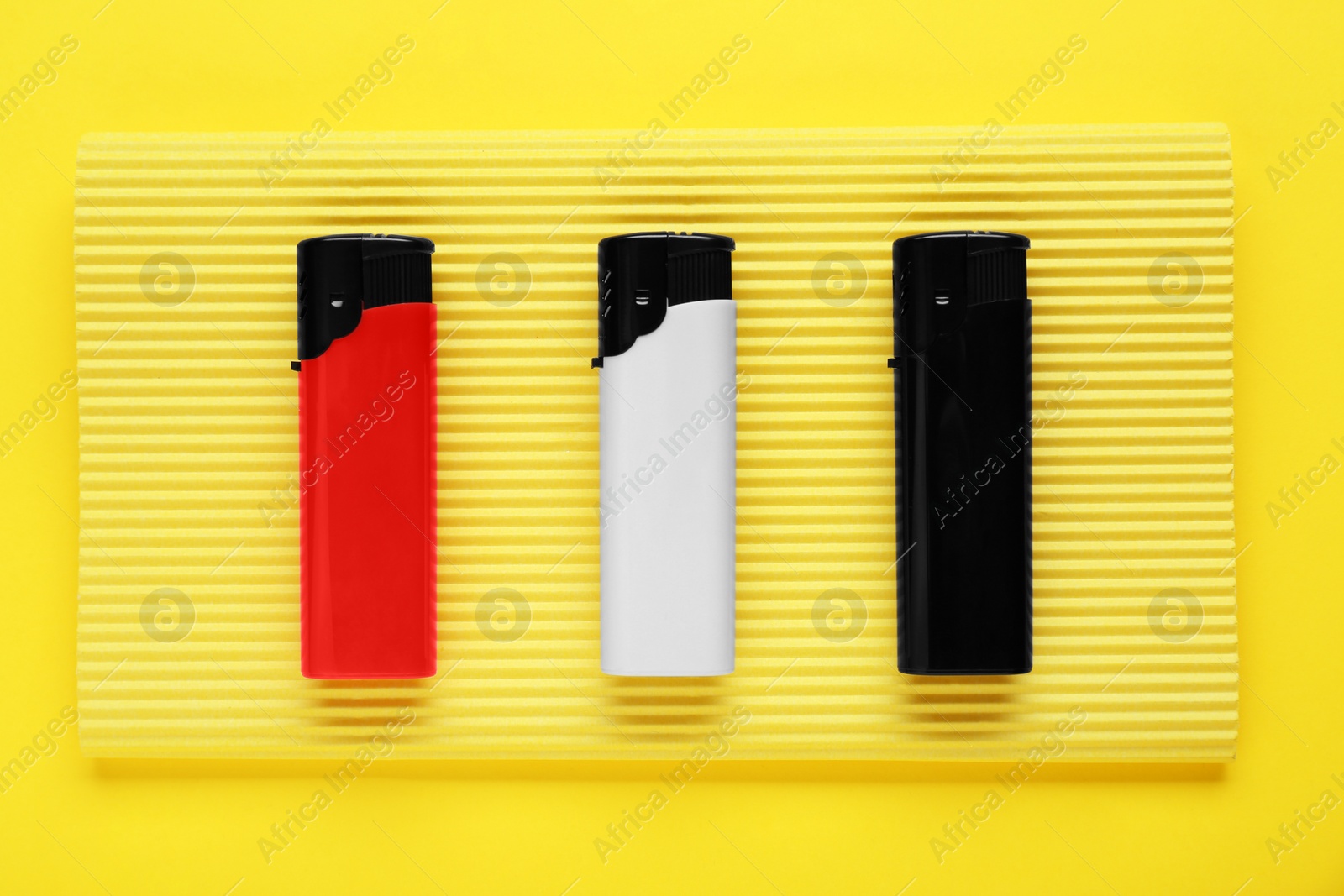 Photo of Stylish small pocket lighters with corrugated fiberboard on yellow background, top view