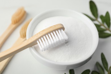 Bamboo toothbrushes, bowl of baking soda and green leaves on white table, closeup