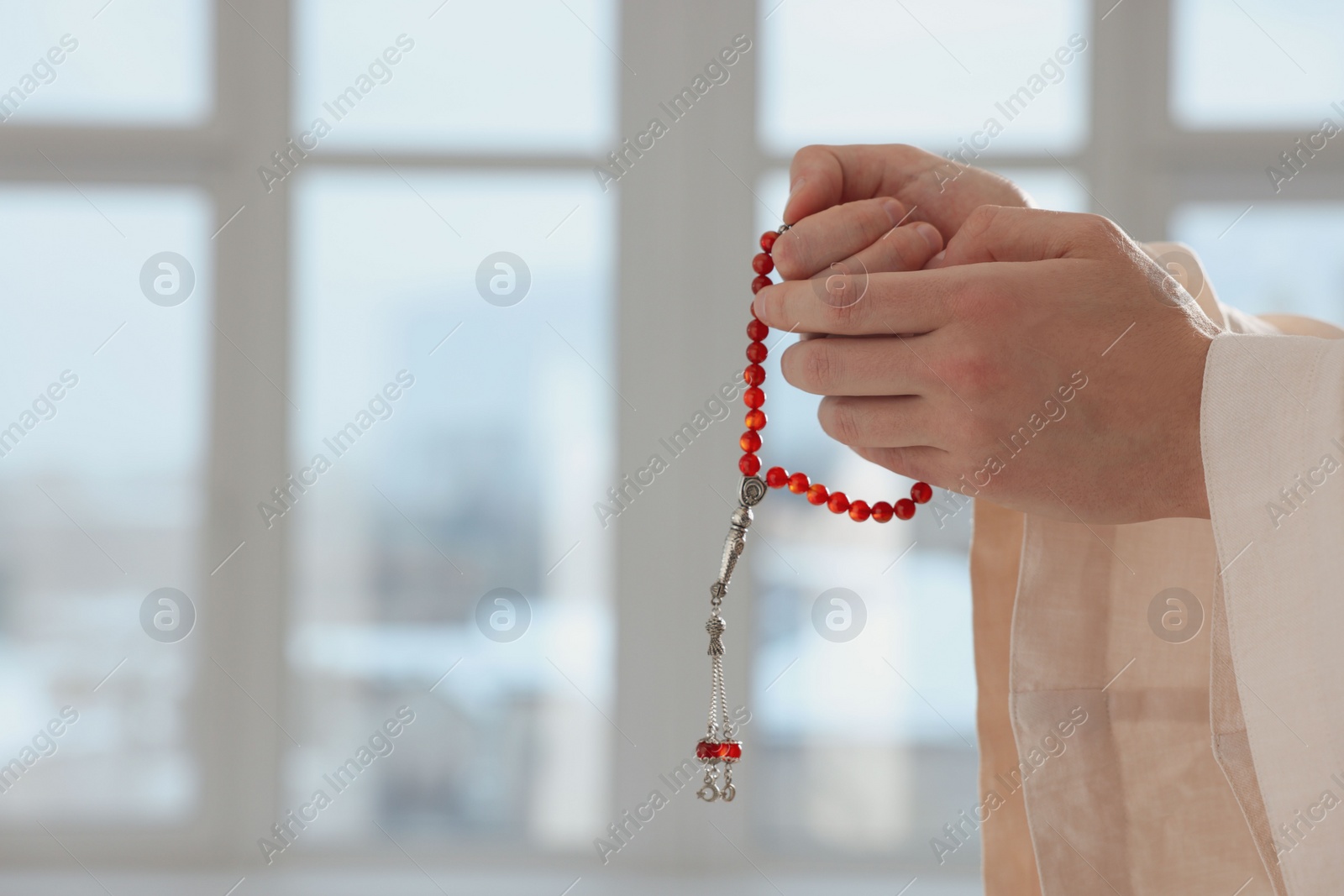 Photo of Muslim man with misbaha praying near window indoors, closeup. Space for text