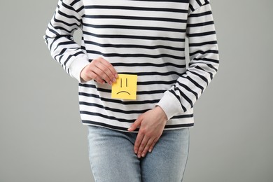 Photo of Woman holding sticky note with drawn sad face and suffering from cystitis on grey background, closeup