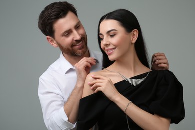 Man putting elegant necklace on beautiful woman against grey background