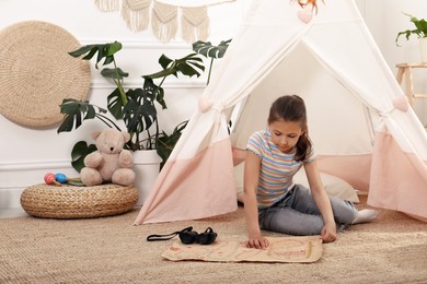 Photo of Cute little girl with treasure map and binoculars near toy wigwam at home