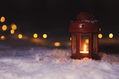 Photo of Lantern with burning candle and Christmas lights on white snow outdoors. Space for text