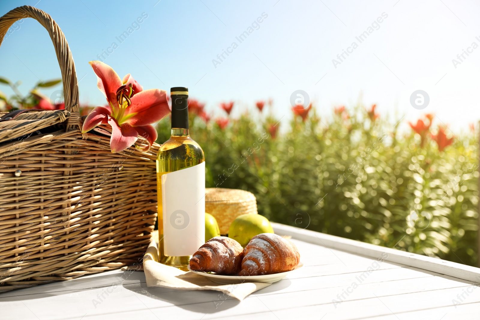 Photo of Composition with picnic basket and bottle of wine on white wooden table in lily field. Space for text