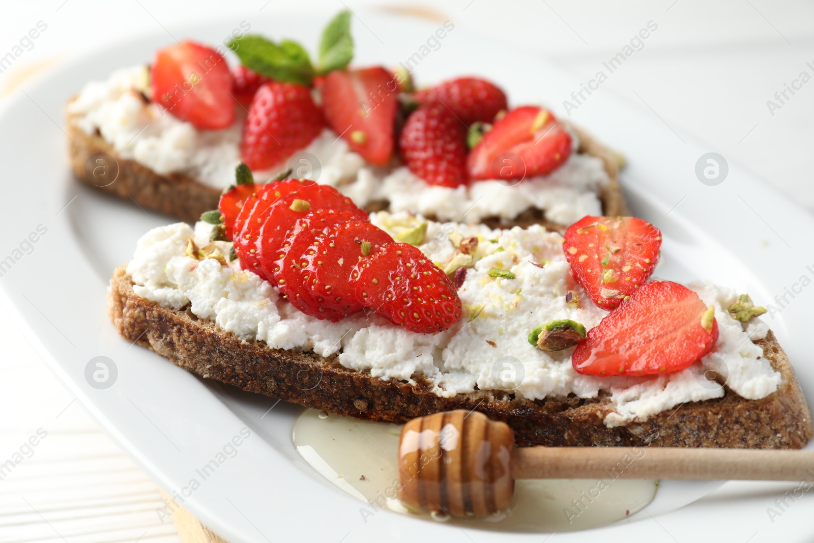 Photo of Delicious ricotta bruschettas with strawberry and pistachios served with honey on white table, closeup