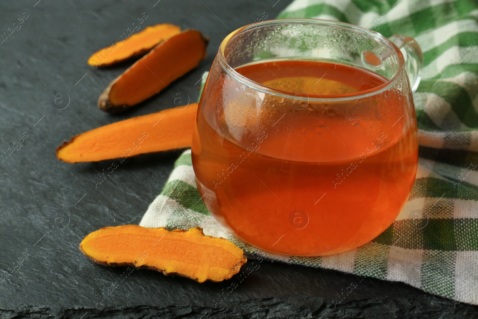 Photo of Glass cup of tasty tea and cut turmeric roots on black textured table, closeup