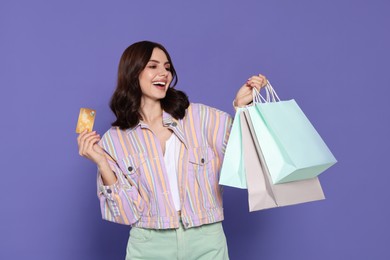 Beautiful young woman with paper shopping bags and credit card on purple background