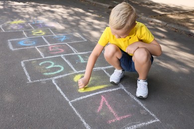 Little boy drawing hopscotch with chalk on asphalt outdoors. Happy childhood