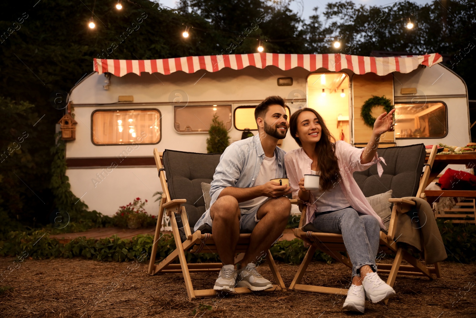 Photo of Happy couple with cups resting near trailer. Camping season