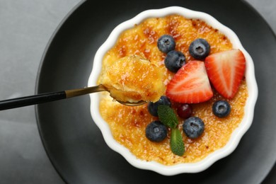 Photo of Taking delicious creme brulee with berries and mint from bowl at grey table, top view