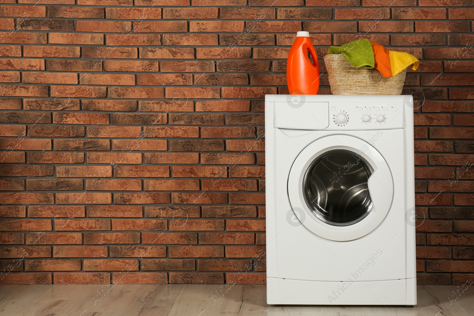 Photo of Modern washing machine with detergent and laundry basket near brick wall. Space for text