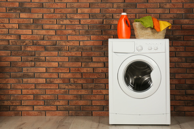 Photo of Modern washing machine with detergent and laundry basket near brick wall. Space for text