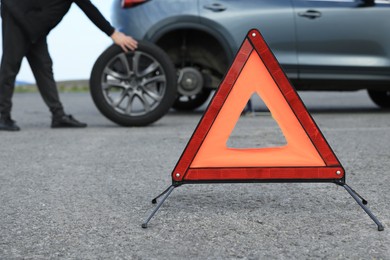 Man changing car tire near road, focus on emergency warning triangle