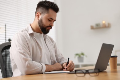 Young man writing down notes during webinar at table in room