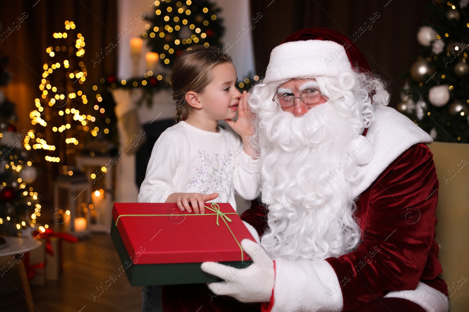 Photo of Little girl whispering in Santa Claus' ear near Christmas tree indoors