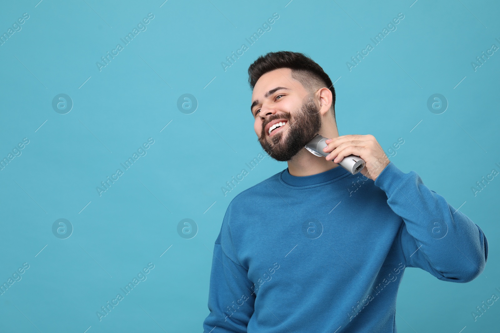 Photo of Handsome young man trimming beard on light blue background. Space for text
