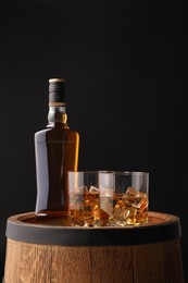 Whiskey with ice cubes in glasses and bottle on wooden barrel against black background