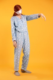 Photo of Young woman wearing pajamas, mask and slippers in sleepwalking state on yellow background