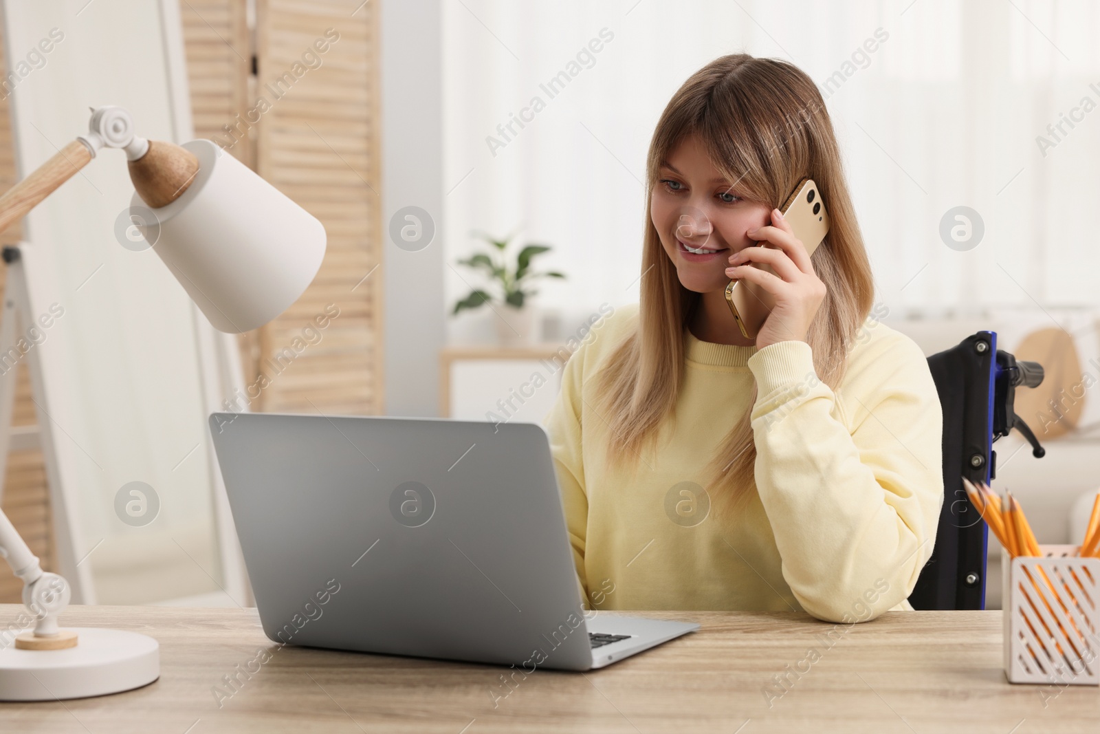 Photo of Woman in wheelchair talking on smartphone while using laptop at table in home office