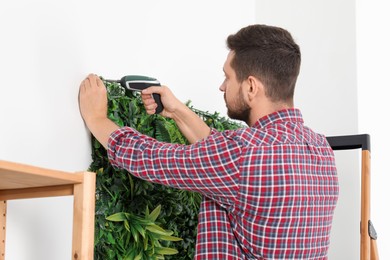 Man with screwdriver installing green artificial plant panel on white wall in room