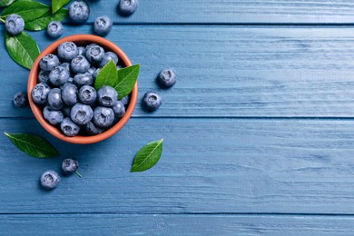 Photo of Tasty fresh blueberries with leaves in bowl on blue wooden table, flat lay. Space for text