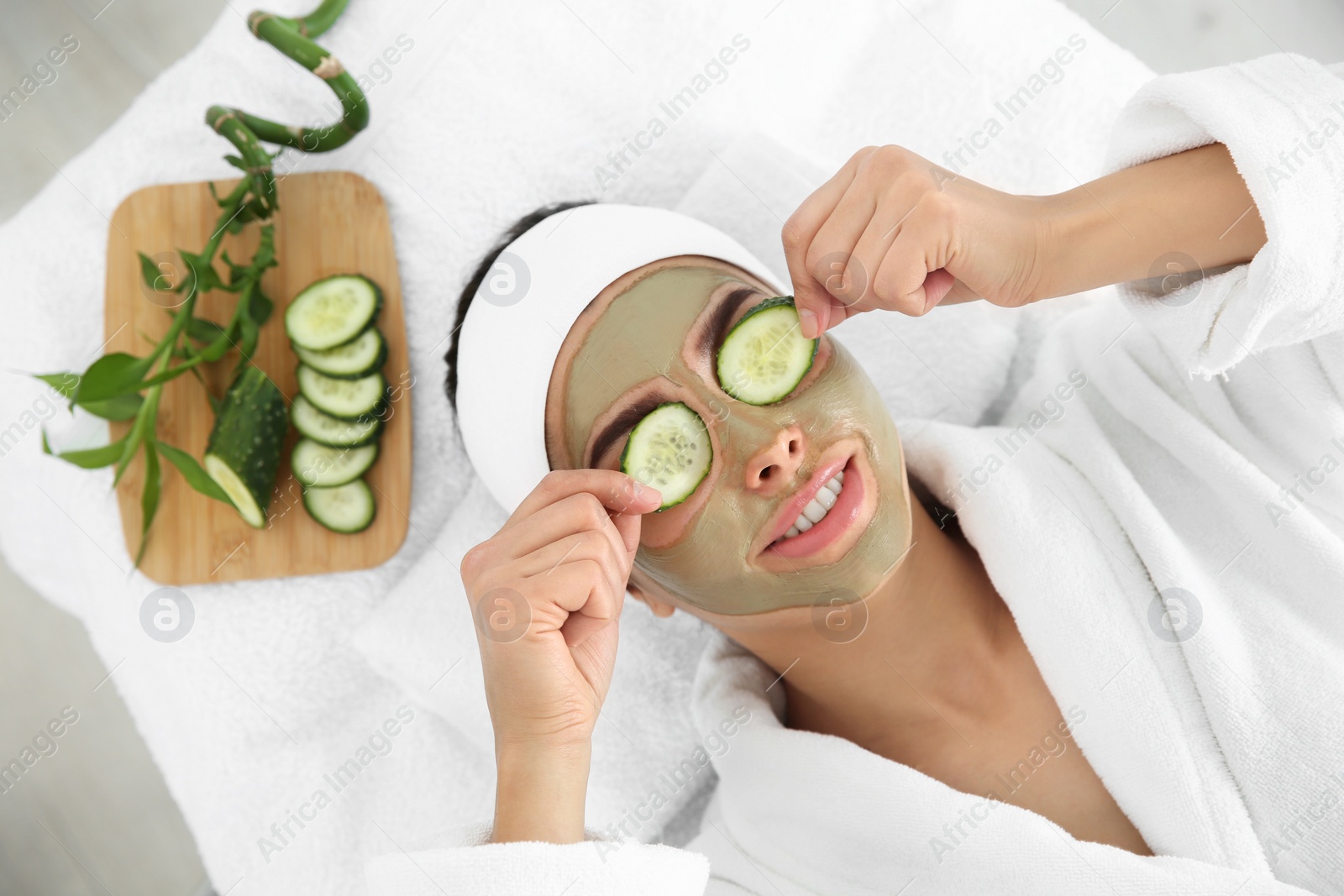 Photo of Pretty woman with clay mask on her face holding cucumber slices in spa salon, above view