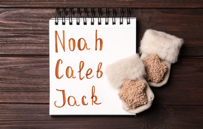 Notebook with different baby names and small mittens on wooden table, flat lay