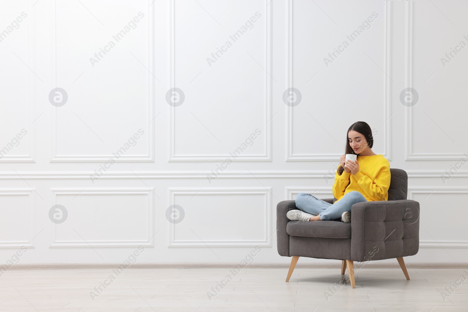 Photo of Beautiful woman with cup of drink sitting in armchair near white wall indoors, space for text