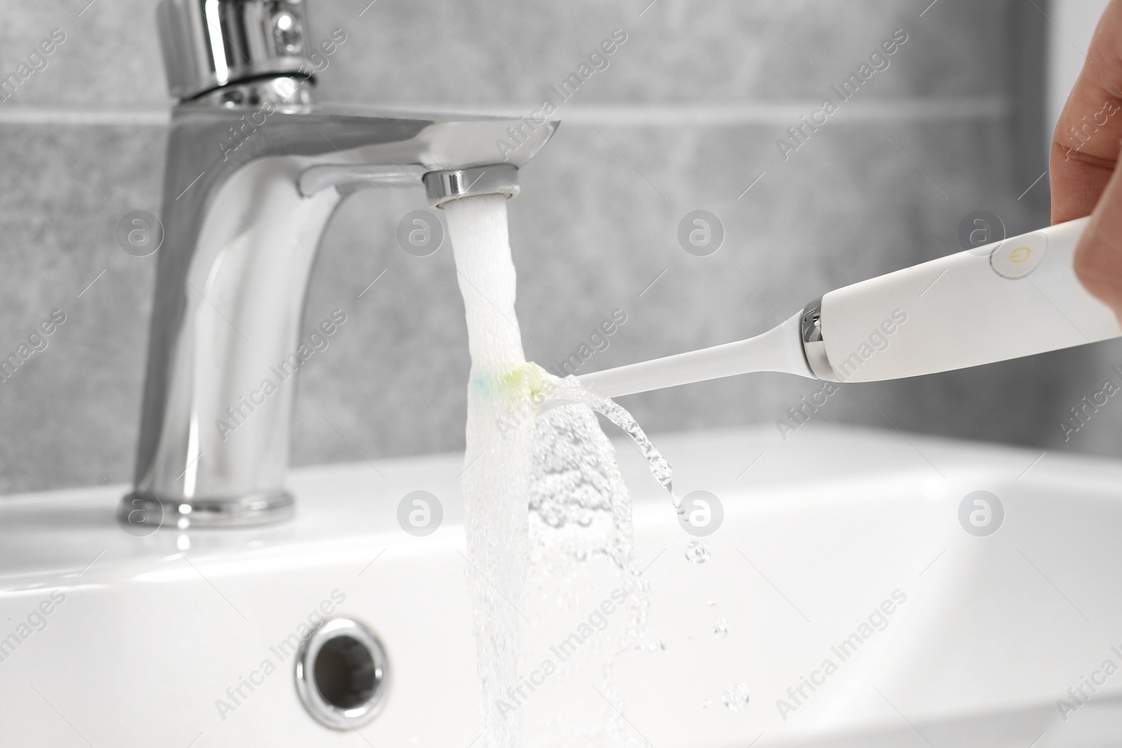 Photo of Man holding electric toothbrush under flowing water above sink in bathroom, closeup