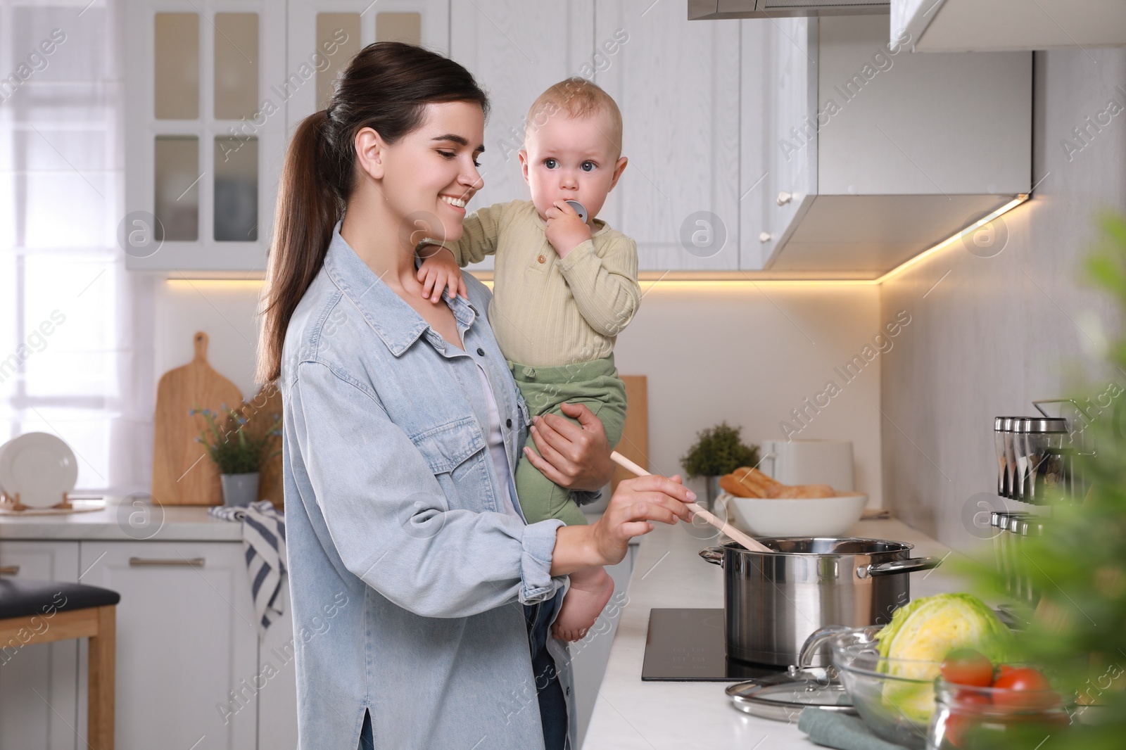 Photo of Happy young woman holding her cute little baby while cooking in kitchen