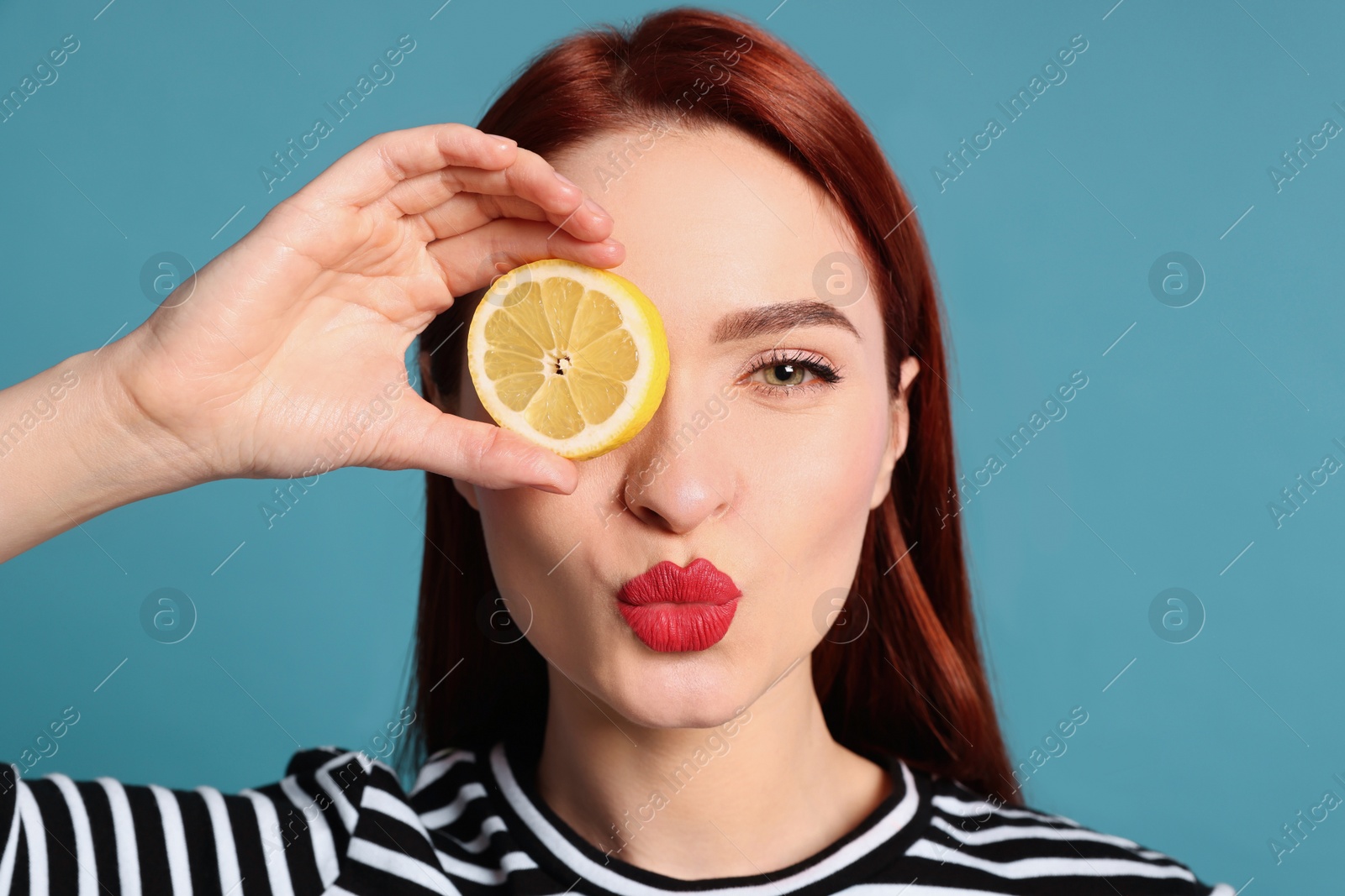 Photo of Beautiful redhead woman covering eye with lemon and blowing kiss on light blue background