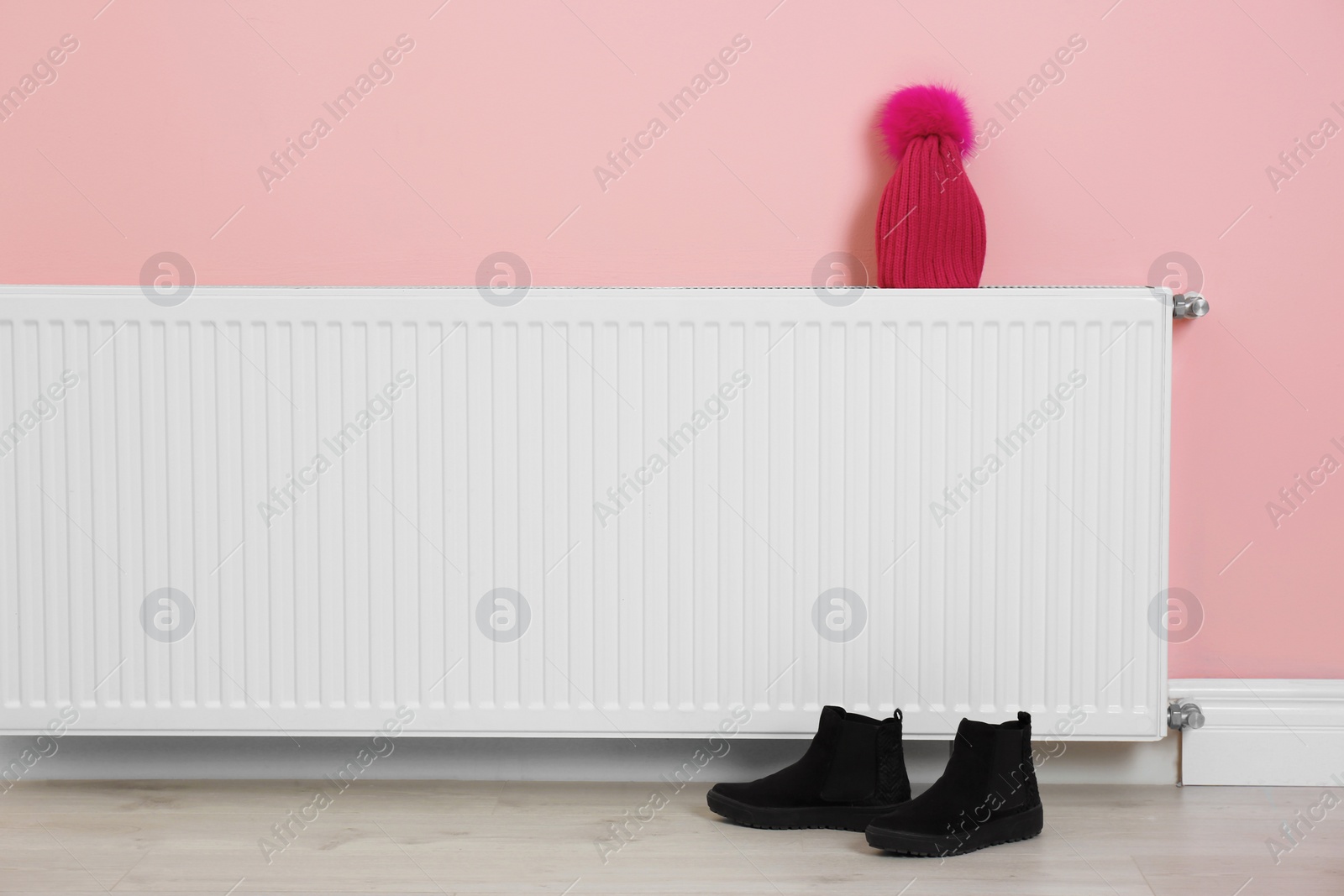 Photo of Heating radiator with knitted cap and shoes near color wall