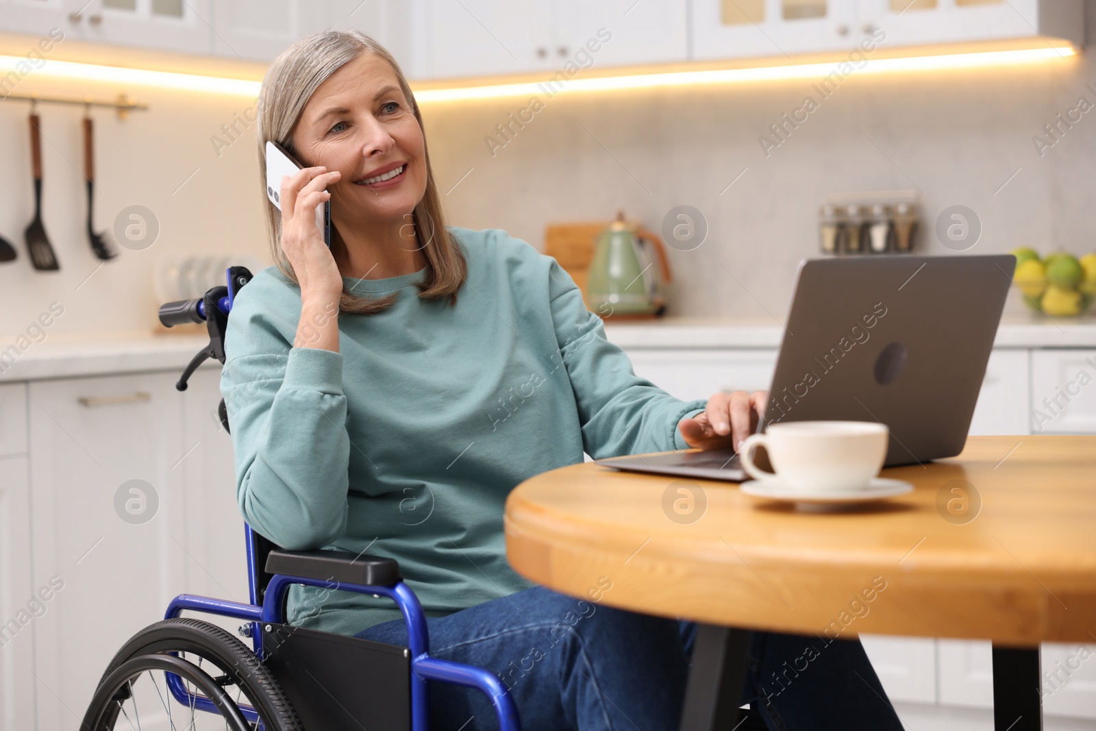 Photo of Woman in wheelchair talking on smartphone while using laptop at home