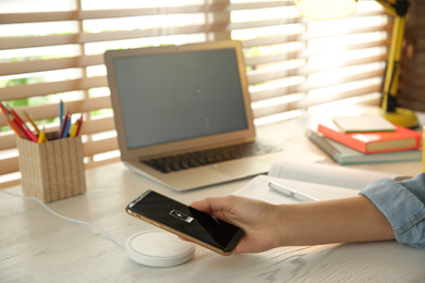 Photo of Woman putting mobile phone onto wireless charger at white wooden table, closeup. Modern workplace accessory