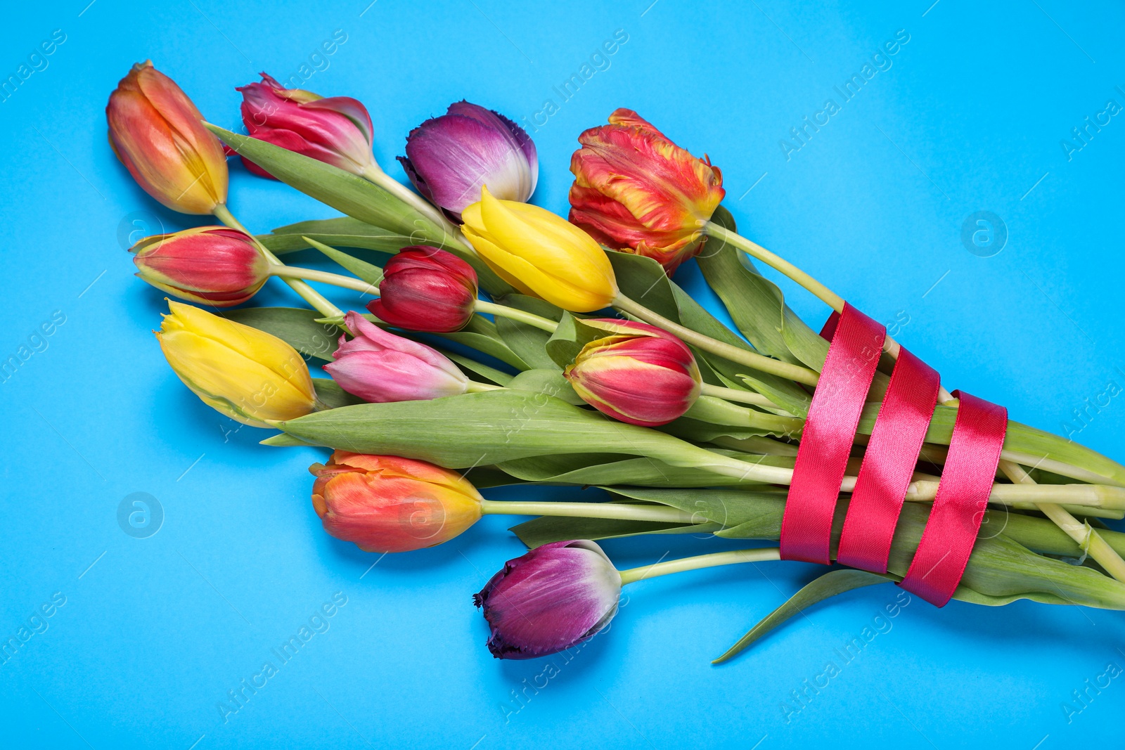 Photo of Bouquet of beautiful colorful tulip flowers tied with red ribbon on light blue background, top view